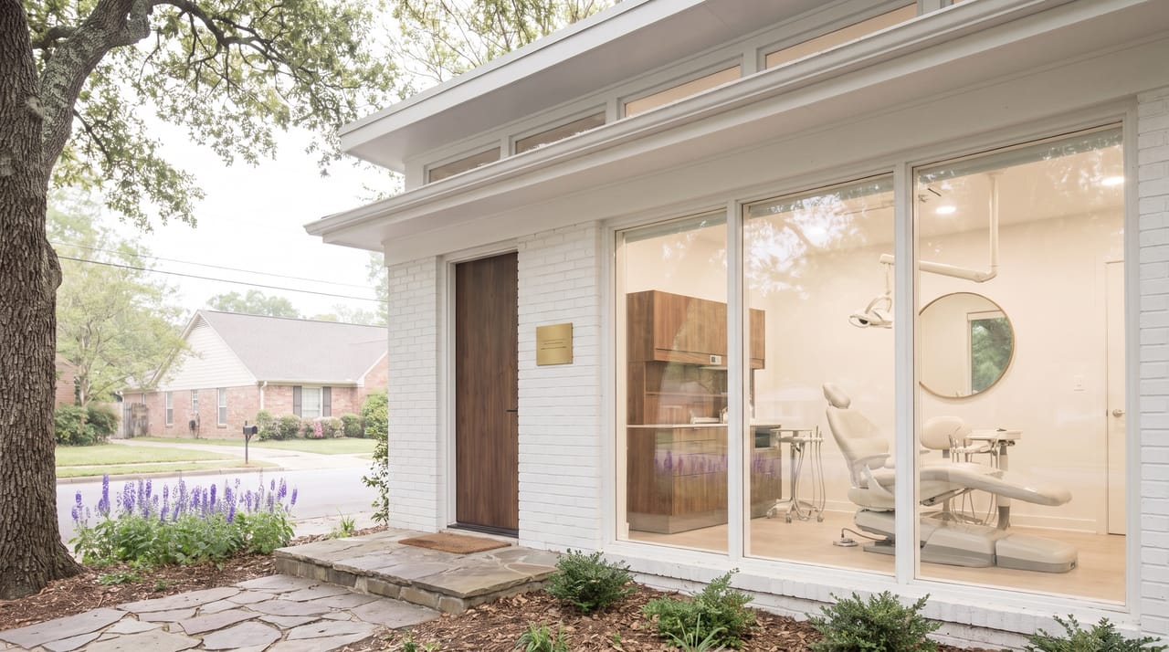 The front of Larkspur Smile Studio on a spring morning: a white brick former portrait studio with a walnut door, a bed of violet larkspur, and a wide window showing a pale grey dental chair, an overhead lamp and a tall round mirror in a bright white room